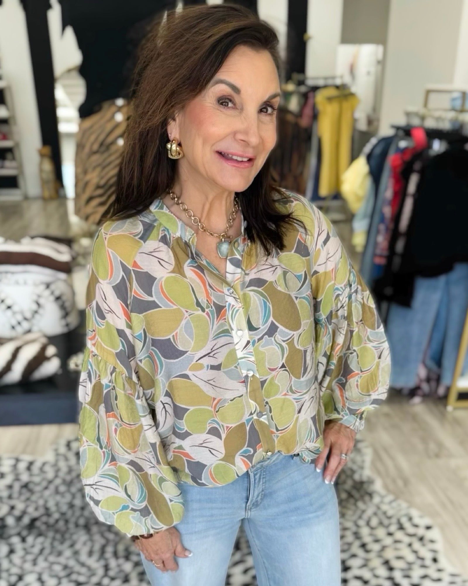 A woman wearing a long-sleeve button-up blouse with a colorful abstract leaf pattern in green, yellow, orange, and blue on a light background, paired with light-wash blue jeans, standing inside a clothing boutique.