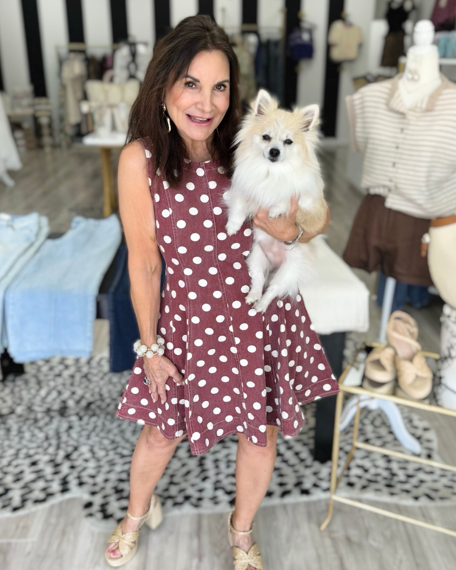 A woman wearing a sleeveless dusty rose pink dress with white polka dots holds a small white dog in a boutique . clothing store. Perfect dress for game day