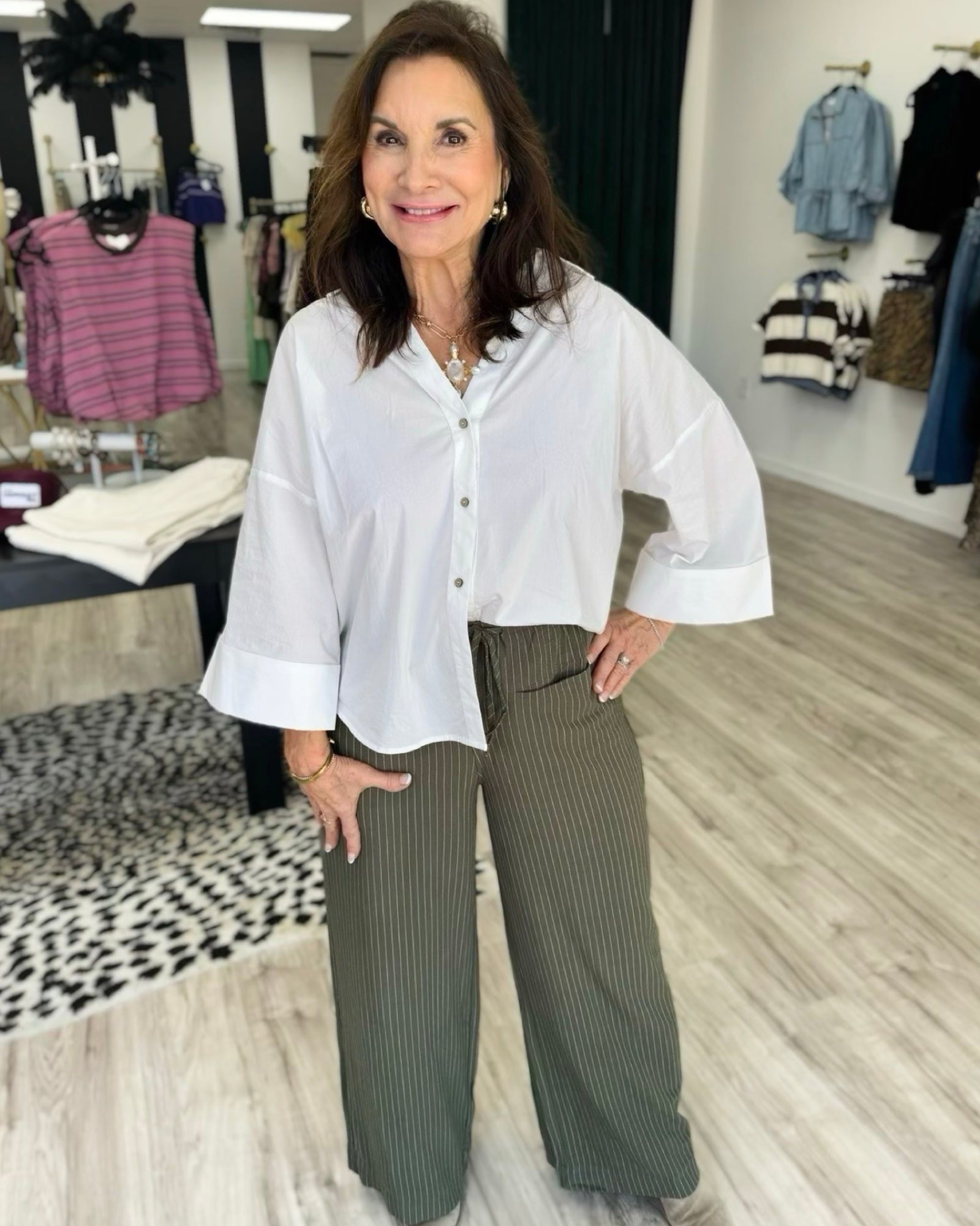 A woman modeling a white button-down shirt with three-quarter sleeves and olive green pinstripe wide-leg pants in a clothing boutique.