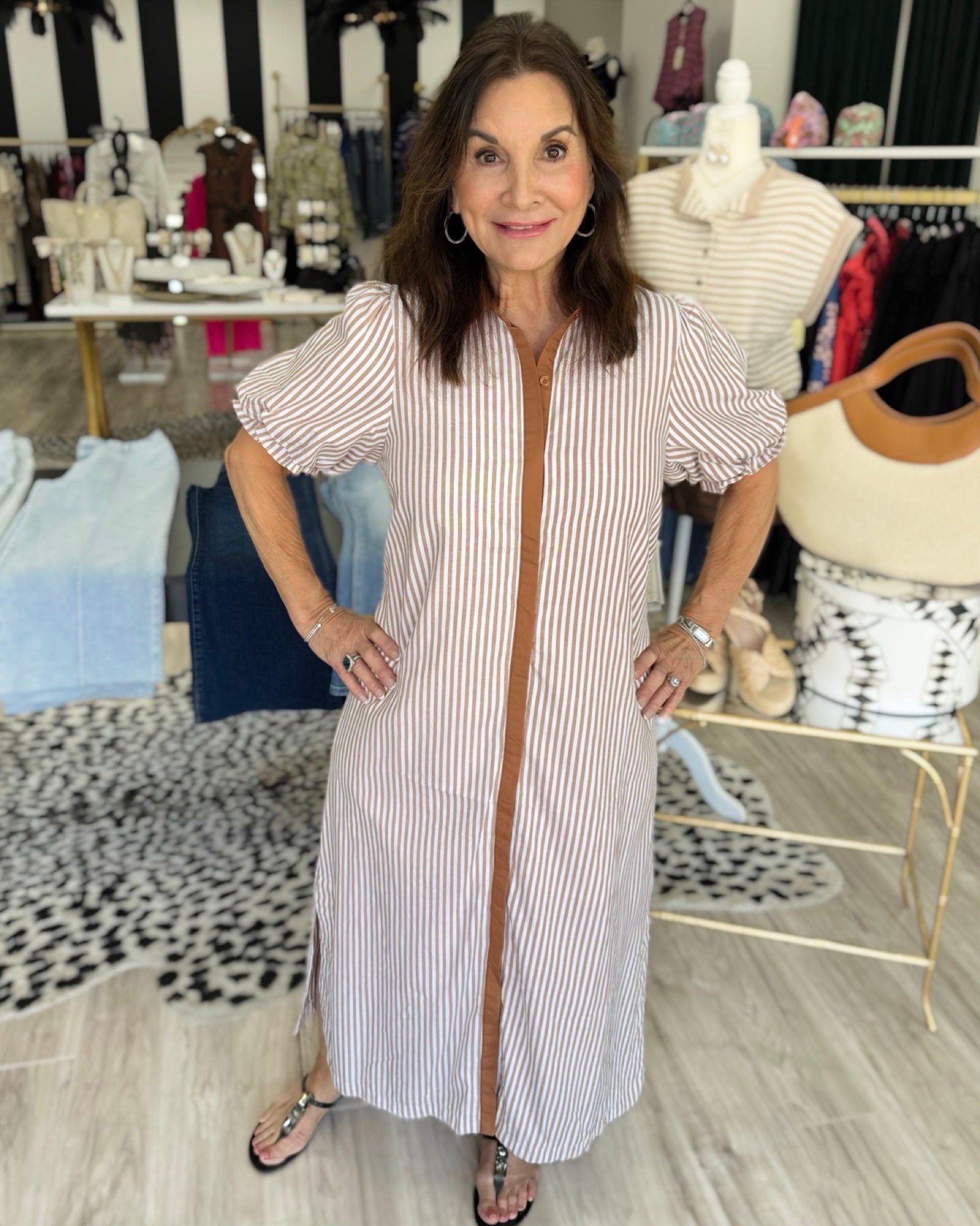 A woman modeling a white and pink vertically striped midi dress with puff sleeves and a brown zipper detail down the front, standing in a boutique setting.