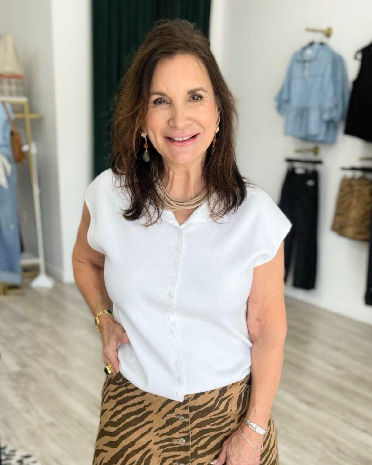 A woman with brown hair is smiling in a boutique, wearing a white short-sleeve button-up blouse and a brown and beige tiger-print skirt. Clothing racks are visible in the softly blurred background.