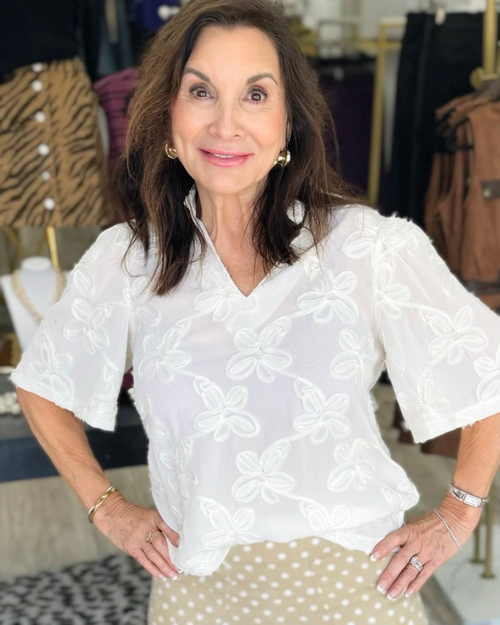 A woman wearing a white short-sleeve blouse with white floral embroidery and a beige polka dot skirt, standing in a clothing store.