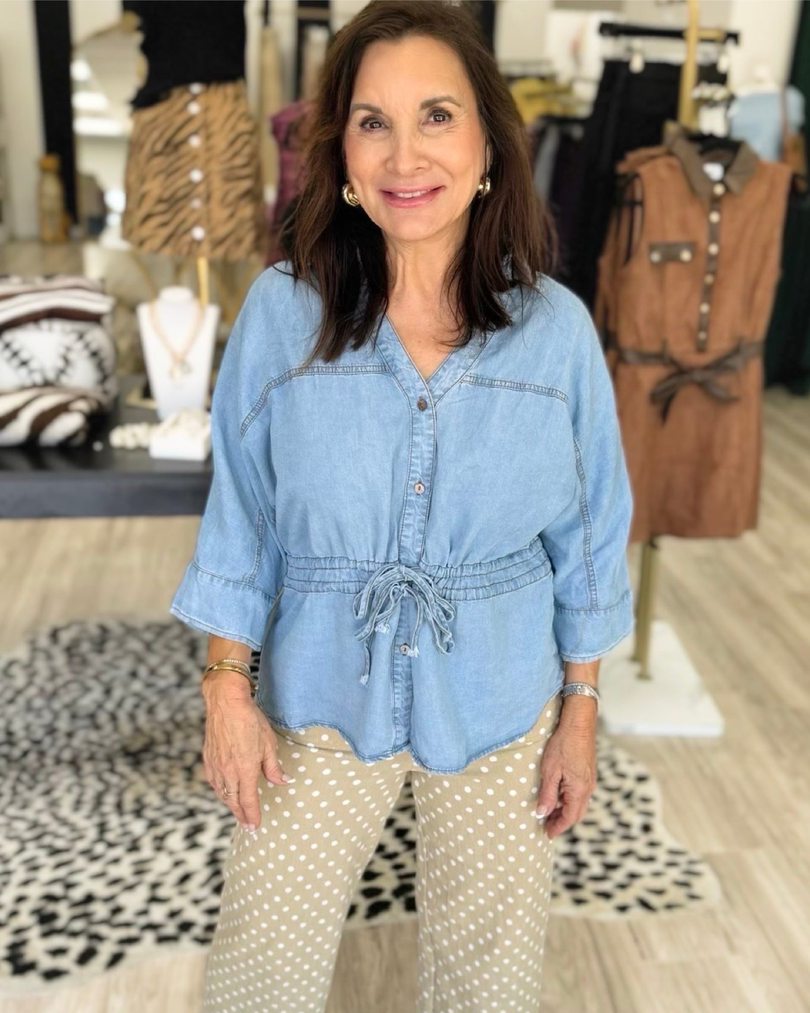 A woman wearing a light blue denim button-up top with a drawstring waist and beige polka-dot pants, standing in a clothing boutique.