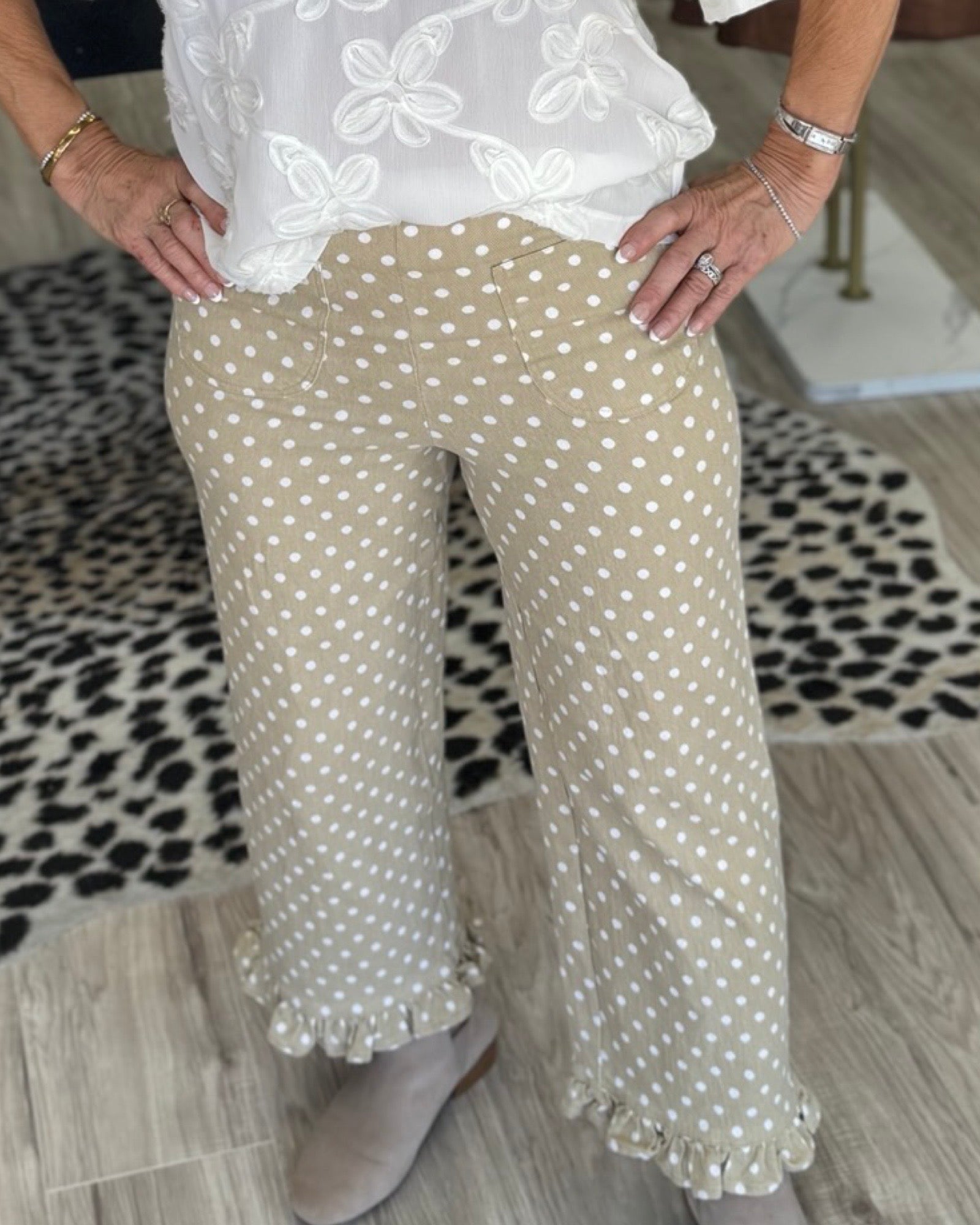 A lady wearing beige wide-leg pants with white polka dots and ruffled hems, paired with a white embroidered top and taupe mule shoes, standing on a wood floor with a black-and-white patterned rug.