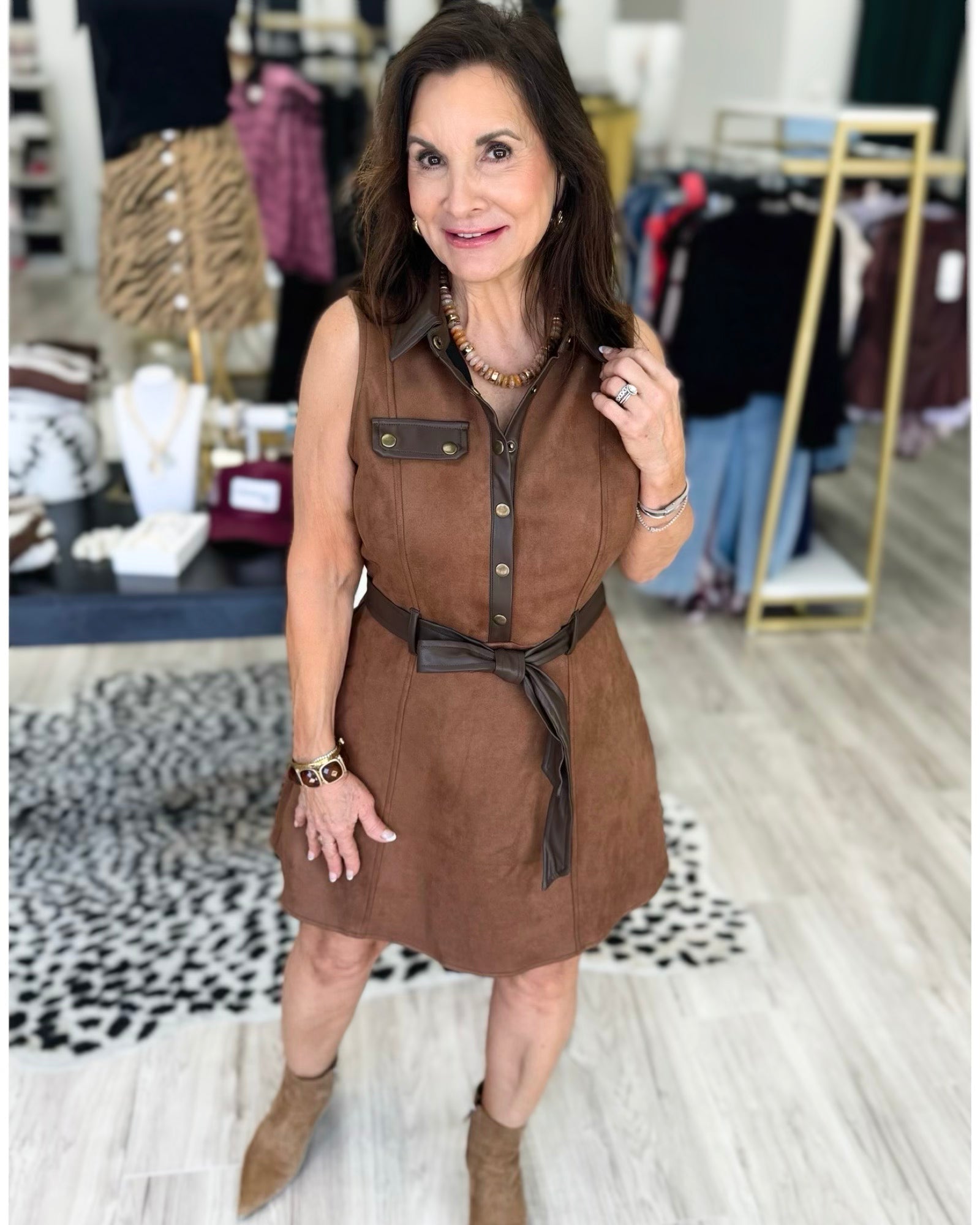 A woman modeling a sleeveless brown faux-suede mini dress with a button-down collar, chest pocket, and matching brown belt, standing in a boutique setting.