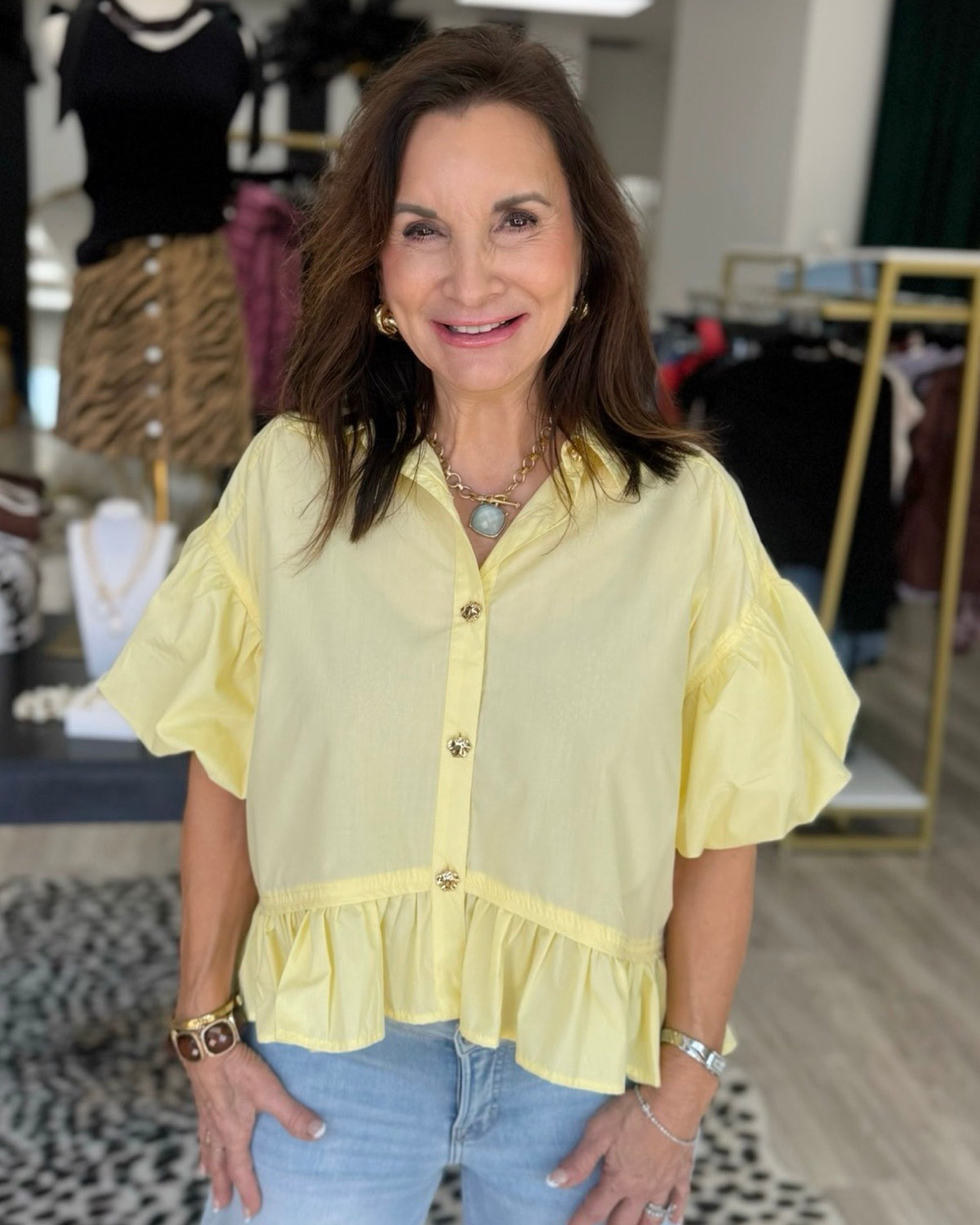 A woman wearing a pale yellow ruffled blouse with gold buttons and light blue jeans, standing in a clothing boutique.